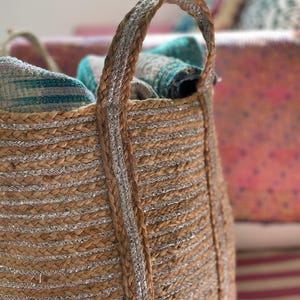 May include: A woven jute basket with silver metallic stripes and handles. The basket is filled with folded textiles in shades of teal, gray, and pink. The basket is designed for storage.