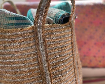 Puede incluir: Una cesta de yute tejida con rayas y asas metálicas plateadas. La cesta está llena de textiles plegados en tonos turquesa, gris y rosa. La cesta está diseñada para el almacenamiento.