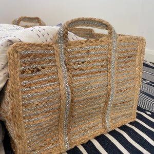 Könnte beinhalten: Eine rechteckige Jute-Tragetasche mit silberfarbenen Metallic-Akzenten. Die Tasche hat zwei Griffe und ist mit einer weiß-lila gemusterten Decke gefüllt. Die Tasche steht auf einem blau-weiß gestreiften Teppich.