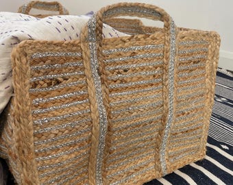 Puede incluir: Un bolso tote rectangular de yute con detalles metálicos plateados. El bolso tiene dos asas y está lleno de una manta blanca y morada estampada. El bolso está sobre una alfombra a rayas azules y blancas.