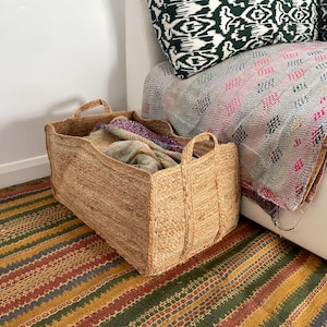 Könnte beinhalten: Ein großer, natürlicher Jutespeicherkorb mit gewebter Textur. Der Korb ist mit einer bunten Decke gefüllt und steht auf einem gestreiften Teppich.