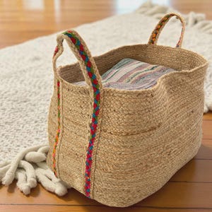 Puede incluir: Una cesta de almacenamiento grande, tejida en yute, con adornos trenzados de colores en las asas y los lados. La cesta está llena de una manta doblada. La cesta está sobre una alfombra de color claro y un suelo de madera.