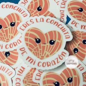 May include: A collection of stickers featuring a cartoon heart-shaped pastry with a smiling face and the text "Eres la conchita de mi corazón" in pink and white lettering. The stickers are on a blue background.