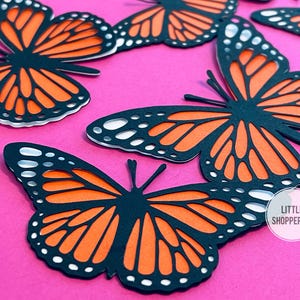 May include: Close-up of several paper monarch butterflies with black outlines and orange wing details, arranged on a bright pink surface. The butterflies have white dots along the edges of their wings. The image includes the text "LITTLE SHOPPERINO".
