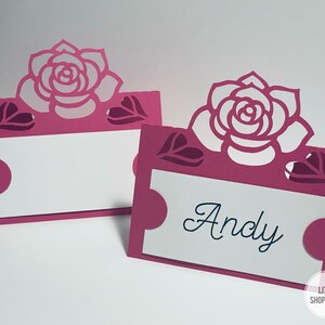 May include: Two pink place card holders with a white rose design and cut-out hearts. The place card on the right says "Andy".