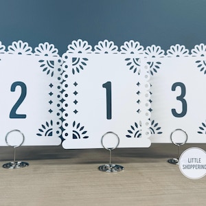 Op de afbeelding: Drie witte tafelnummerkaarten met decoratieve randen en de nummers 1, 2 en 3 in een vet lettertype. Elke kaart wordt vastgehouden door een zilveren draadhouder. De kaarten staan op een lichtbruin oppervlak tegen een donkerblauwe achtergrond.