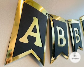 Pode incluir: Faixa preta e dourada com as letras "ABBY" em letras douradas em bandeiras pretas. As bandeiras são conectadas por um cordão dourado.