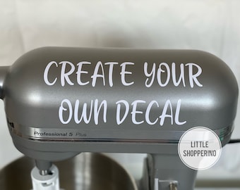 Puede incluir: Una batidora de pie plateada con una calcomanía blanca que dice "CREATE YOUR OWN DECAL" y una pegatina que dice "LITTLE SHOPPERINO".