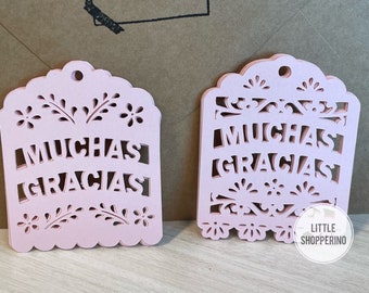 May include: Two pink paper cut-out decorations with the text "MUCHAS GRACIAS" in a decorative font. The decorations are shaped like a traditional Mexican paper lantern.