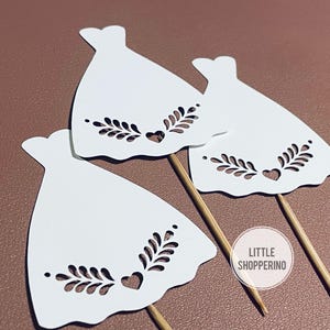 Op de afbeelding: Drie witte papieren jurkuitsparingen met decoratieve blad- en hartdetails, bevestigd aan houten spiesjes. De uitsparingen zijn gerangschikt op een bruin oppervlak. De afbeelding bevat een cirkelvormig logo met de tekst "LITTLE SHOPPERINO".