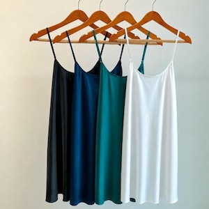 May include: Four sleeveless slip dresses with thin spaghetti straps, each hanging on a wooden hanger. The dresses are presented in a row, featuring a smooth, satin-like finish in solid colours of black, navy blue, teal green, and crisp white.