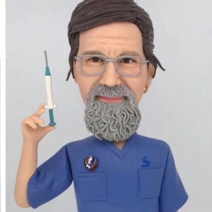 May include: A custom bobblehead figurine depicting a person with a gray beard and glasses, wearing a blue scrub top with a circular pin and blue pants. The figure holds a small syringe in one hand and stands on a gray circular base labeled Greatful Dead.