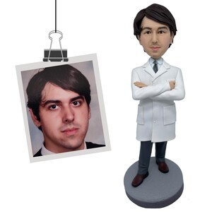 May include: A custom bobblehead figurine depicting a person with dark hair wearing a white lab coat, a light blue collared shirt, a dark tie, and dark trousers, standing on a round gray base next to a photograph of the same person clipped to a wire.