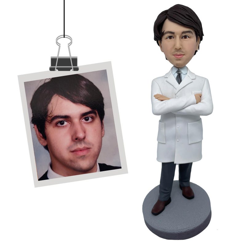 May include: A custom bobblehead figurine depicting a person with dark hair wearing a white lab coat, a light blue collared shirt, a dark tie, and dark trousers, standing on a round gray base next to a photograph of the same person clipped to a wire.