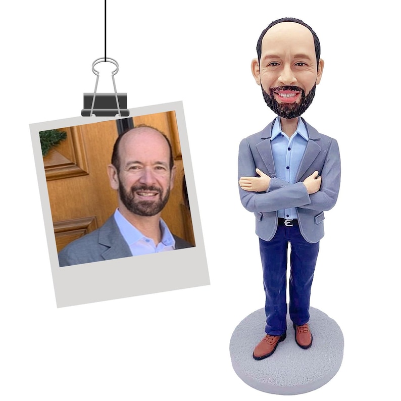 Puede incluir: Figura de bobblehead de un hombre con blazer gris, camisa azul y pantal&oacute;n azul, con los brazos cruzados. Una foto de un hombre con blazer gris est&aacute; sujeta en la parte superior. El bobblehead se encuentra sobre una base circular gris.