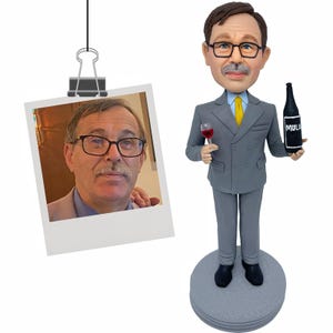 Puede incluir: Figura de un hombre con traje gris sosteniendo una copa de vino y una botella de vino negra. La figura tiene bigote y gafas. Una foto de un hombre con gafas y bigote est&aacute; sujeta a la imagen.