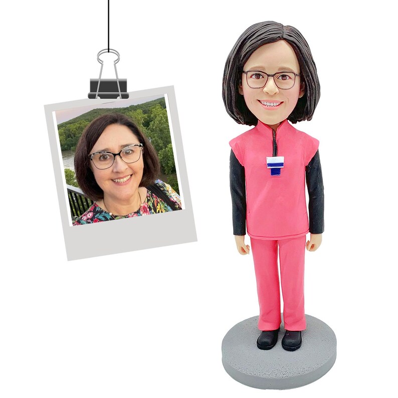 May include: A custom bobblehead figurine featuring a person with dark shoulder-length hair and glasses, wearing a pink scrub top over a black long-sleeved shirt and pink pants, standing on a gray circular base next to a photo of the same person.