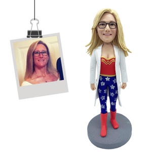 May include: A custom bobblehead figurine featuring a person with blonde hair and black-rimmed glasses, wearing a white lab coat over a red top with a gold emblem and blue leggings with white stars, standing on a gray circular base next to a photo.
