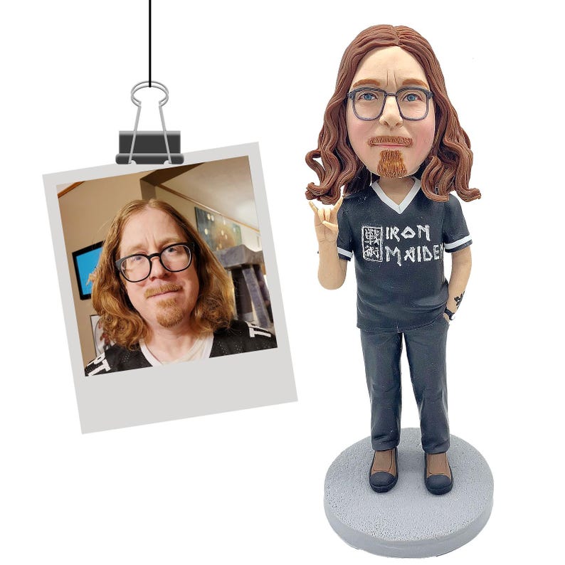 Puede incluir: Una figura de bobblehead personalizada de una persona con cabello largo y ondulado color casta&ntilde;o rojizo, gafas y perilla, que lleva una camiseta negra con "IRON MAIDEN" impresa. La figura est&aacute; sobre una base circular gris. Una foto est&aacute; sujeta en la parte superior.