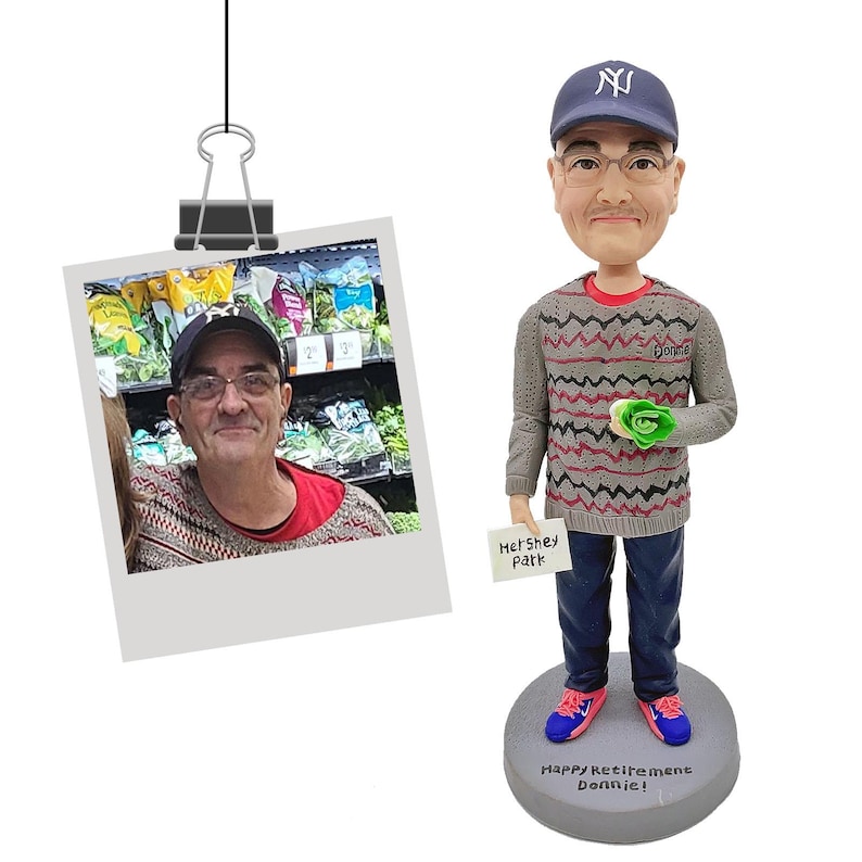 Puede incluir: Figura de bobblehead de un hombre con gorra de b&eacute;isbol de los Yankees de Nueva York, gafas y un su&eacute;ter gris. La figura sostiene un objeto verde y un letrero que dice "Hershey Park". Se adjunta una foto del hombre.