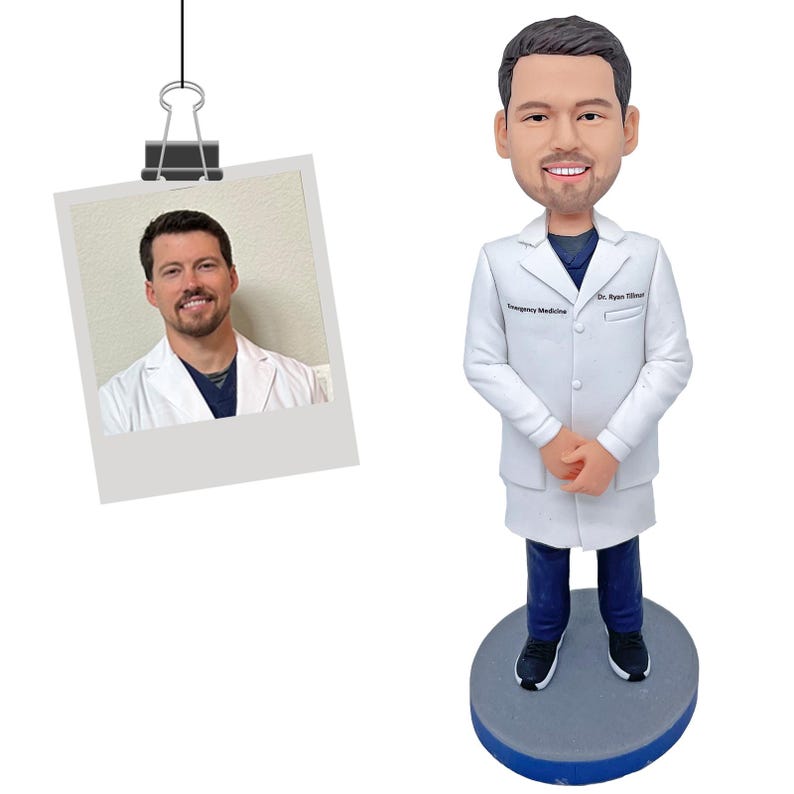 May include: A custom bobblehead figurine depicting a medical professional with brown hair and a beard, wearing a white lab coat over a dark blue shirt and pants. The coat features the text Dr. Ryan Tillman and Emergency Medicine.