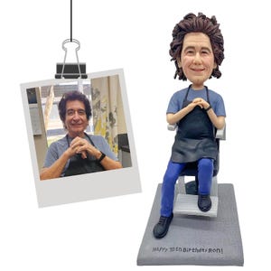 Puede incluir: Figura de un hombre en un sill&oacute;n de barbero, con camisa azul, delantal negro y pantal&oacute;n azul. Se adjunta una foto del hombre. La base dice "Happy 70th Birthday Ron!"