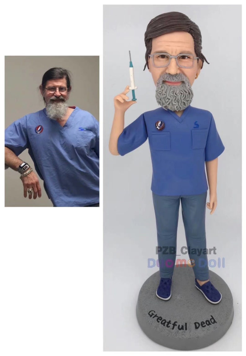 May include: A custom bobblehead figurine depicting a person with a gray beard and glasses, wearing a blue scrub top with a circular pin and blue pants. The figure holds a small syringe in one hand and stands on a gray circular base labeled Greatful Dead.