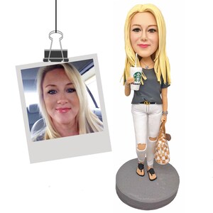 Puede incluir: Una figura de bobblehead personalizada de una mujer con cabello rubio largo, que lleva una camiseta gris, jeans blancos rasgados y sandalias negras. Sostiene una taza de Starbucks y un bolso tote a cuadros. Una foto Polaroid est&aacute; adjunta.