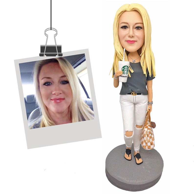 Puede incluir: Una figura de bobblehead personalizada de una mujer con cabello rubio largo, que lleva una camiseta gris, jeans blancos rasgados y sandalias negras. Sostiene una taza de Starbucks y un bolso tote a cuadros. Una foto Polaroid est&aacute; adjunta.