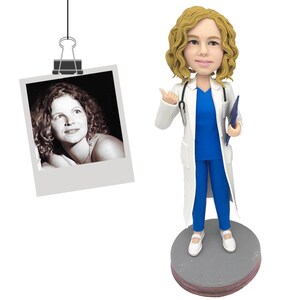 May include: A custom bobblehead figurine of a medical professional with curly blonde hair, wearing a white lab coat over a blue scrub top and pants, holding a blue clipboard, with a stethoscope around the neck, standing on a gray circular base.