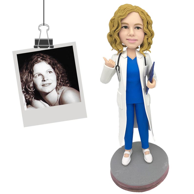 May include: A custom bobblehead figurine of a medical professional with curly blonde hair, wearing a white lab coat over a blue scrub top and pants, holding a blue clipboard, with a stethoscope around the neck, standing on a gray circular base.