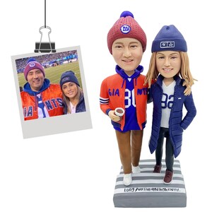 Puede incluir: Una figura de dos personas con ropa de los New York Giants, incluyendo una chaqueta roja y azul y un gorro azul. Se adjunta una foto de la pareja con ropa de los Giants. La base de la figura tiene el texto "Long you live and high you fly..."