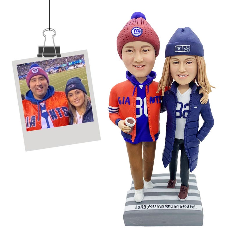 Puede incluir: Una figura de dos personas con ropa de los New York Giants, incluyendo una chaqueta roja y azul y un gorro azul. Se adjunta una foto de la pareja con ropa de los Giants. La base de la figura tiene el texto "Long you live and high you fly..."