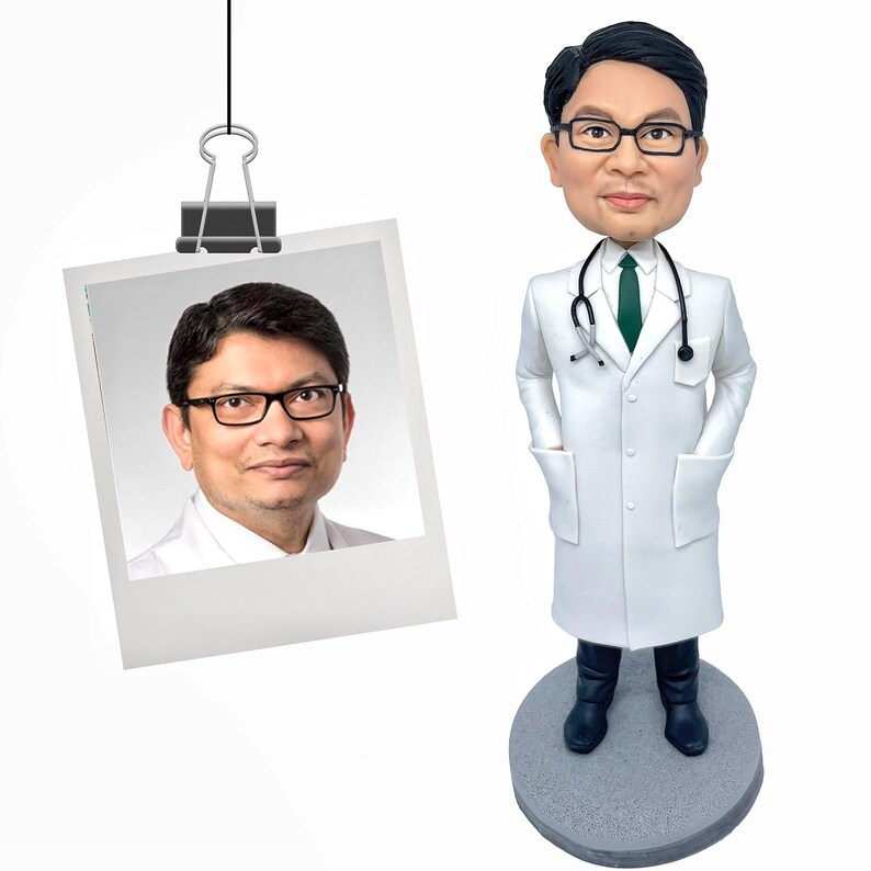 May include: A custom bobblehead figurine of a doctor wearing a white lab coat, a green necktie, and black-rimmed glasses with a stethoscope around the neck, standing on a circular gray base next to a photograph of the person depicted.