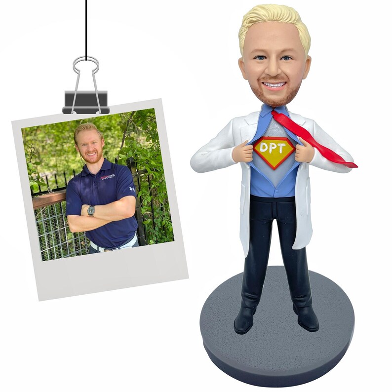 May include: A custom bobblehead figure of a person with blonde hair, wearing a white lab coat over a blue shirt and dark trousers, pulling open the coat to reveal a yellow and red superhero chest emblem with the letters DPT, standing on a base.