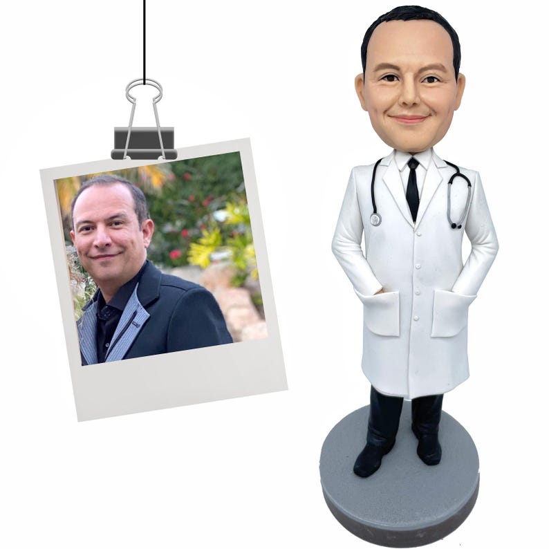 May include: A custom bobblehead figure of a person with short dark hair, wearing a white lab coat, a black tie, and dark trousers, standing on a circular gray base. A stethoscope is draped around the neck of the figure, which has hands in pockets.