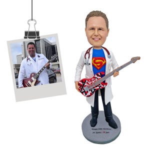 May include: A custom bobblehead figure of a person wearing a white lab coat over a blue shirt with a red and yellow Superman logo, holding a red, white, and black electric guitar. A stethoscope is around the neck. The base reads Happy 50th Birthday, Dr. Love - jme.