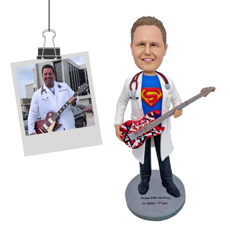 May include: A custom bobblehead figure of a person wearing a white lab coat over a blue shirt with a red and yellow Superman logo, holding a red, white, and black electric guitar. A stethoscope is around the neck. The base reads Happy 50th Birthday, Dr. Love - jme.