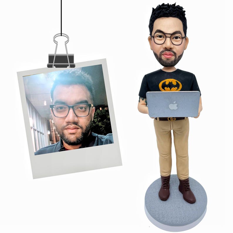 Puede incluir: Una figura de bobblehead personalizada de un hombre sosteniendo una computadora port&aacute;til, vistiendo una camiseta negra con un s&iacute;mbolo de murci&eacute;lago amarillo, pantalones color caqui y botas marrones. Una foto Polaroid del hombre cuelga cerca.