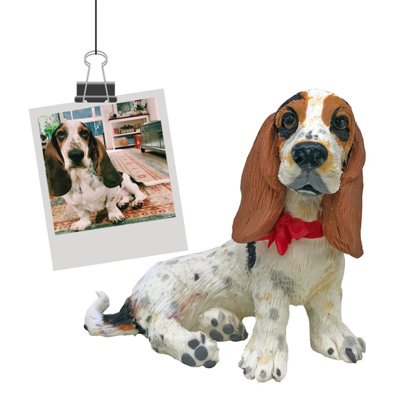 Puede incluir: Figura detallada de un perro Basset Hound, con cuerpo blanco con manchas negras y marrones, largas orejas marrones y un lazo rojo. Una foto polaroid de un Basset Hound est&aacute; sujeta en la parte superior.