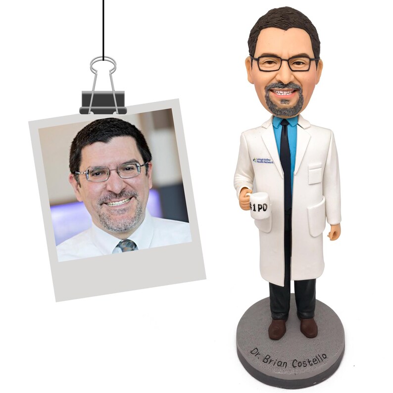 May include: A custom bobblehead figure of a person with dark hair, a beard, and glasses, wearing a white lab coat, blue shirt, and black tie. The figure holds a white mug labeled #1 PD and stands on a gray base inscribed with Dr. Brian Costello.