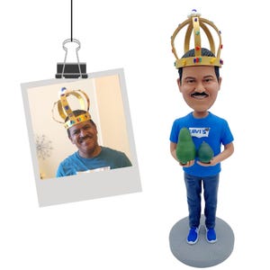 Puede incluir: Una figura de un hombre con una corona dorada y una camiseta azul con el logo de Levi's, sosteniendo dos aguacates verdes. La figura est&aacute; sobre una base gris. Se adjunta una foto de un hombre con una corona.