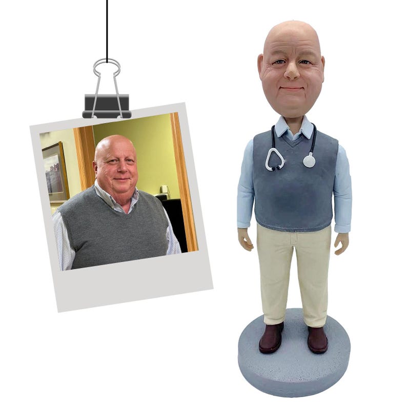 May include: A custom bobblehead figurine depicting a bald person with a light complexion, wearing a light blue collared shirt under a grey sweater vest, beige trousers, and dark shoes, with a stethoscope draped around the neck on a round base.