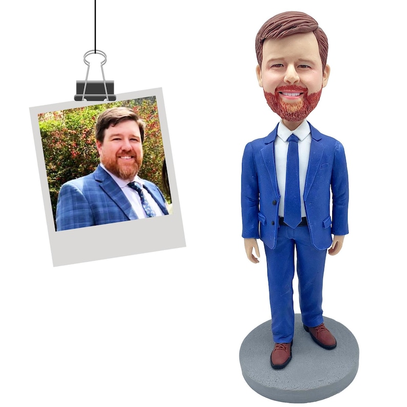 Puede incluir: Una figura de bobblehead personalizada de un hombre con un traje azul, camisa blanca y corbata azul, de pie sobre una base circular gris. Una foto del hombre con un traje azul est&aacute; sujeta en la parte superior. La figura tiene cabello casta&ntilde;o y barba roja.