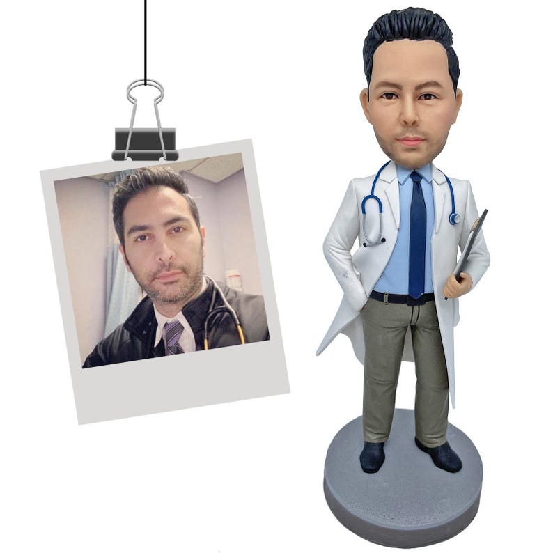 May include: A custom bobblehead figurine of a doctor wearing a white lab coat, light blue shirt, dark blue tie, and khaki trousers. The figure holds a clipboard and has a stethoscope around the neck, standing on a round gray base next to a photo.