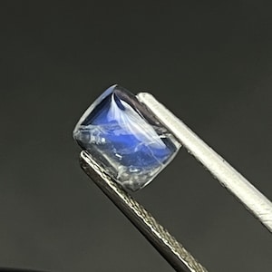 May include: A blue moonstone gemstone with a rectangular cut, held in tweezers.