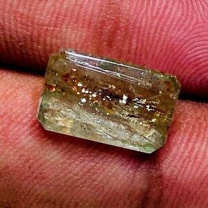 Pode incluir: Uma gema verde e marrom com formato retangular. A gema tem uma aparência clara e vítrea e está engastada em uma configuração simples e sem adornos.