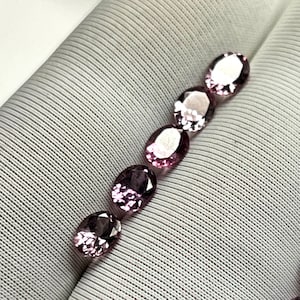 Puede incluir: Una colección de ocho gemas ovaladas en tonos de rosa y morado. Las gemas están dispuestas en dos filas sobre una superficie de tela blanca.