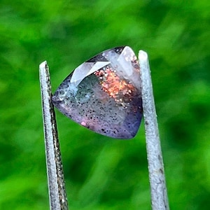 Op de afbeelding: Een driehoekige edelsteen die door een pincet wordt vastgehouden. De edelsteen heeft een mix van kleuren, waaronder paars, blauw en oranje, met een gespikkeld patroon. De achtergrond is een wazig groen.