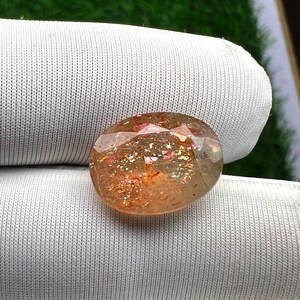 Pode incluir: Uma grande gema de sunstone, de forma oval, de cor marrom avermelhada e com brilho iridescente. A pedra está colocada sobre um fundo branco texturizado.